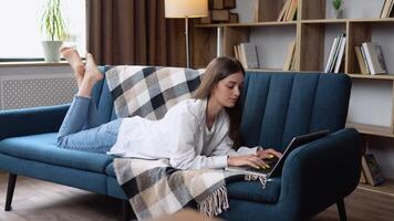 Young woman lying on her stomach a sofa using a laptop computer to surf the internet with a happy relaxed smile video