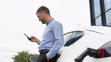 joven hermoso europeo hombre con inalámbrico auriculares, utilizando teléfono mientras propensión en su eléctrico auto, cargando el batería a ciudad poder estación video