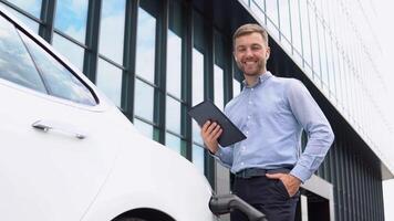 joven europeo empresario con inalámbrico auriculares y tableta cerca eléctrico auto, cargando el batería a ciudad poder estación video