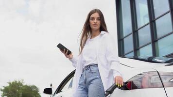 joven mujer es en pie cerca el eléctrico coche y mira a el inteligente teléfono. eléctrico cargando estación video