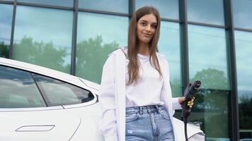 Smiling young caucasian girl plugging electricity cable in electric vehicle for charging on sunny mall parking, selective focus. Lifestyle and ecology concept video