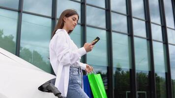 jovem menina depois de comprando produtos com uma compras saco é em pé perto a cobrando elétrico carro video