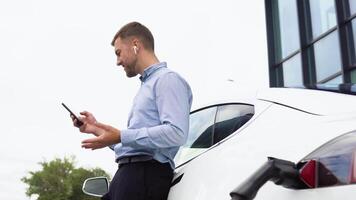 joven hermoso europeo hombre con inalámbrico auriculares, Hablando en un charla en su teléfono inteligente mientras propensión en su eléctrico auto, cargando el batería a ciudad poder estación video