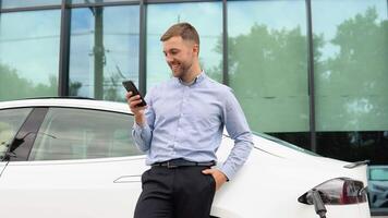 Portrait of young handsome european man, speaking on a chat on his smartphone while leaning on his electric car, charging the battery at city power station video