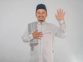 Moslem Asian man looking to the camera with happy face expression while waving his hand to greet others photo