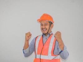 asiático hombre trabajador vistiendo la seguridad casco mira contento celebrando su victoria por apretando su puños en contra gris antecedentes foto