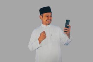 Moslem Asian man clenched fist showing excitement when looking to his mobile phone photo