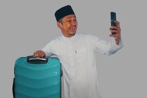 Moslem Asian man carrying suitcase showing happy expression when looking his phone photo