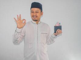 adulto musulmán asiático hombre sonriente y dar Okay dedo firmar mientras participación dinero foto