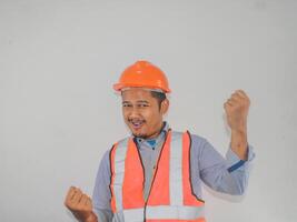 asiático hombre trabajador vistiendo la seguridad casco mira contento celebrando su victoria por apretando su puños en contra foto