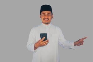 musulmán asiático hombre sonriente contento mientras participación móvil teléfono y señalando a el Derecha lado foto