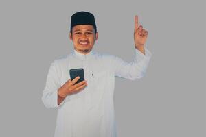 Moslem Asian man smiling and pointing finger up while holding a smartphone photo