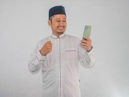 Moslem Asian man clenched fist showing excitement while looking to his mobile phone photo