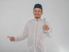 Moslem man smiling while holding a bottle of drinking water and pointing to the right side photo