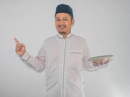 Moslem Asian man smiling and pointing to the right side while holding empty dinner plate photo