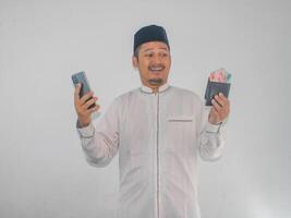 asiático musulmán hombre sonriente contento mientras participación móvil teléfono y demostración papel dinero desde su billetera foto