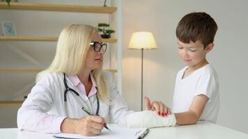 sorridente anziano femmina famiglia medico nel uniforme consulenza poco ragazzo paziente con un' rotto mano a verifica incontro nel clinica video