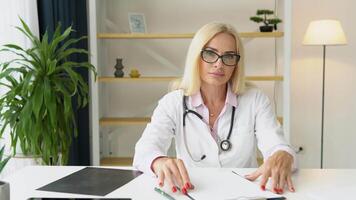 senior europeisk kvinna läkare bär vit medicinsk täcka och stetoskop ser på kamera video