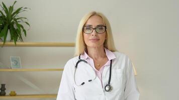 Happy senior woman doctor wearing white medical coat and stethoscope looking at camera. Positive general practitioner close up face portrait. Smiling female physician posing in hospital office video