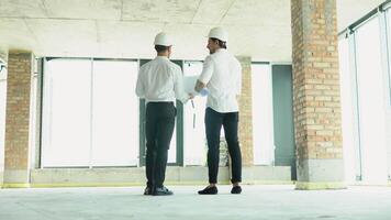 A young engineers in safety helmets standing on an under-construction building holding a blueprint of the structure and working together video