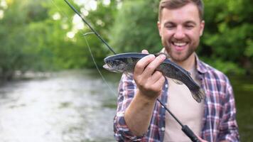 Fischer ruht auf das Fluss und fängt Forelle, lächelt und zeigt an das Fisch im das Kamera video