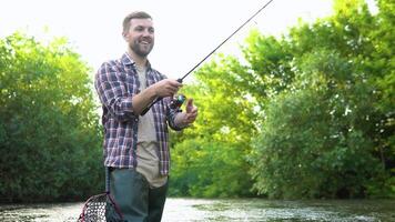 een visser gooit een visvangst hengel staand kniediep in de water van de rivier. vlieg visvangst voor forel. zomer vakantie en mensen concept video