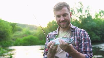 Fisherman rests on the river and catches trout, smiles and shows the fish in the camera video