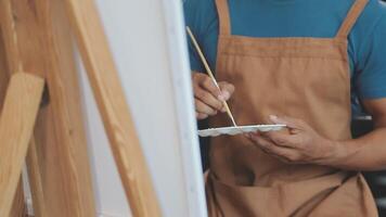 Over the shoulder selective focus shot of male artist using brush while adding color paint to pencil drawing on canvas video