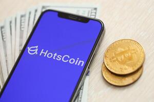 KYIV, UKRAINE - MARCH 15, 2024 Hotscoin logo on iPhone display screen with dollars and bitcoins on table photo