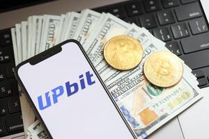 KYIV, UKRAINE - MARCH 15, 2024 Upbit logo on iPhone display screen with dollars and bitcoins on MacBook photo