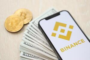 KYIV, UKRAINE - MARCH 15, 2024 Binance logo on iPhone display screen with dollars and bitcoins on table photo