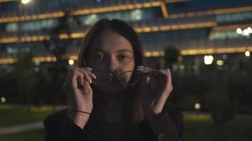 en contra el fondo de un moderno ciudad a noche, con sus brillante luces esclarecedor el horizonte, un joven exitoso mujer de negocios pone en su lentes y con confianza miradas dentro el cámara. cerca arriba. lento movimiento. 4k video