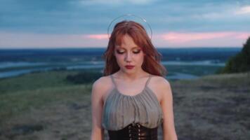 magnifique Jeune femme avec inhabituel maquillage et une semblable à un halo accessoire prend une étape vers l'avant et regards dans le caméra. sa rouge cheveux est doucement ébouriffé par le brise. portrait est ensemble dans nature, dans le soir. proche en haut. lent mouvement. 4k video