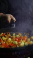 Chef of the restaurant is actively mixing colorful vegetables in a black frying pan. Oil is flying spectacularly from the pan in all directions, creating a beautiful scene against the black background. Vertical. Close up. 4k. High angle video