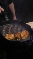 Juicy chicken meat, with a golden crust, is stirred by the chef in a black griddle pan, with splashes of boiling oil flying everywhere. Vertical. Close up. 4k. High angle video