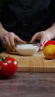 cucinare è spingendo un' piatto pieno con bianca salsa in direzione il telecamera, contro un' fondale di fresco, vivace verdure per un' insalata ricetta. lui è vestito nel un' nero uniforme contro un' buio sfondo. verticale. vicino su. 4k. video