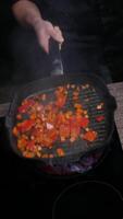 Cook mixes diced carrots and bell peppers, lifting the pan over a preheated black induction hob. Vertical. Close up. 4k. High angle video