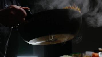 Cook holds a large frying pan in his hands, mixing vegetables and meat together as he throws them into the air against a dark abstract background. Close up. 4k. video