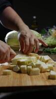 Man is dicing a zucchini with a knife, surrounded by vegetables, on a black backdrop. Vertical. Close up. 4k. video