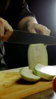 large green zucchini is being sliced by a chef using a large kitchen knife against an abstract black backdrop. Vertical. Close up. 4k. video