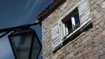 A lantern in front of the house. Creative. A large stone house taken at an angle which has a window on which small flowers grow, a lantern and a roof can be seen video