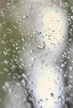 A photo of rain drops on the window glass with a blurred view of the blossoming green trees. Abstract image showing cloudy and rainy weather conditions