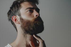 ai generado hermoso joven barbado hombre es acuerdo mano en barba y mirando lejos en un gris antecedentes foto