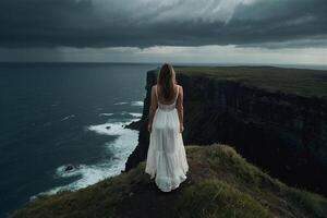 un mujer en un blanco vestir soportes en el borde de un acantilado con vista a el Oceano foto