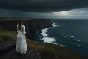 a woman in a white dress stands on a cliff overlooking the ocean photo