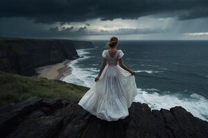 a woman in a white dress stands on a cliff overlooking the ocean photo