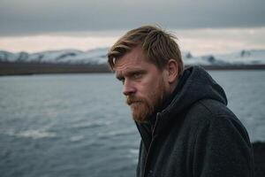 a man with a beard and a hoodie standing near the water photo