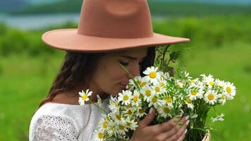 Chamomile woman. Happy woman in a white dress and brown hat holds a bouquet of daisies in her hands and smells them video