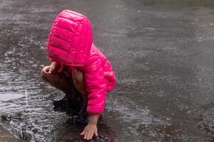 un asiático pequeño niña vistiendo un rosado chaqueta y jugando en el lluvia en el asfalto la carretera foto