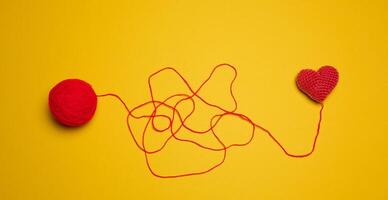 A ball of thread and a knitted red heart on a yellow background, top view photo